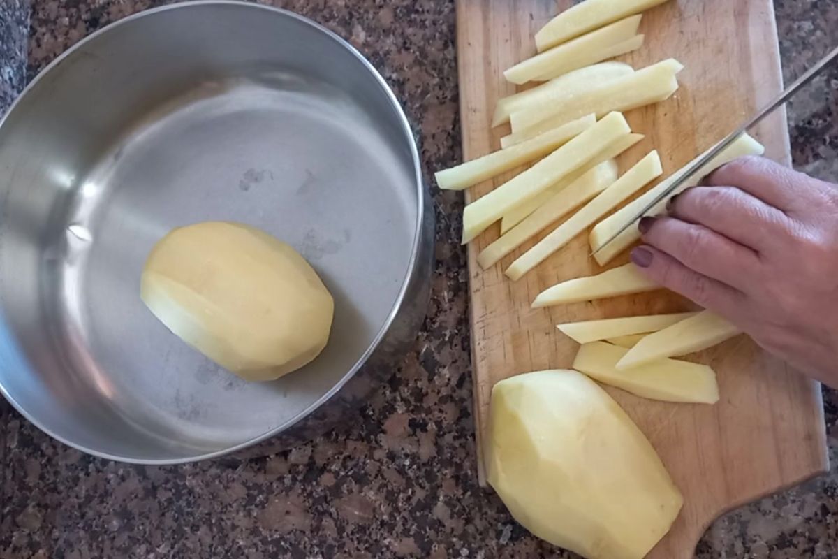 Batata inglesa crua sendo cortada em palitos sobre tábua de madeira, etapa de preparo da batata frita na airfryer