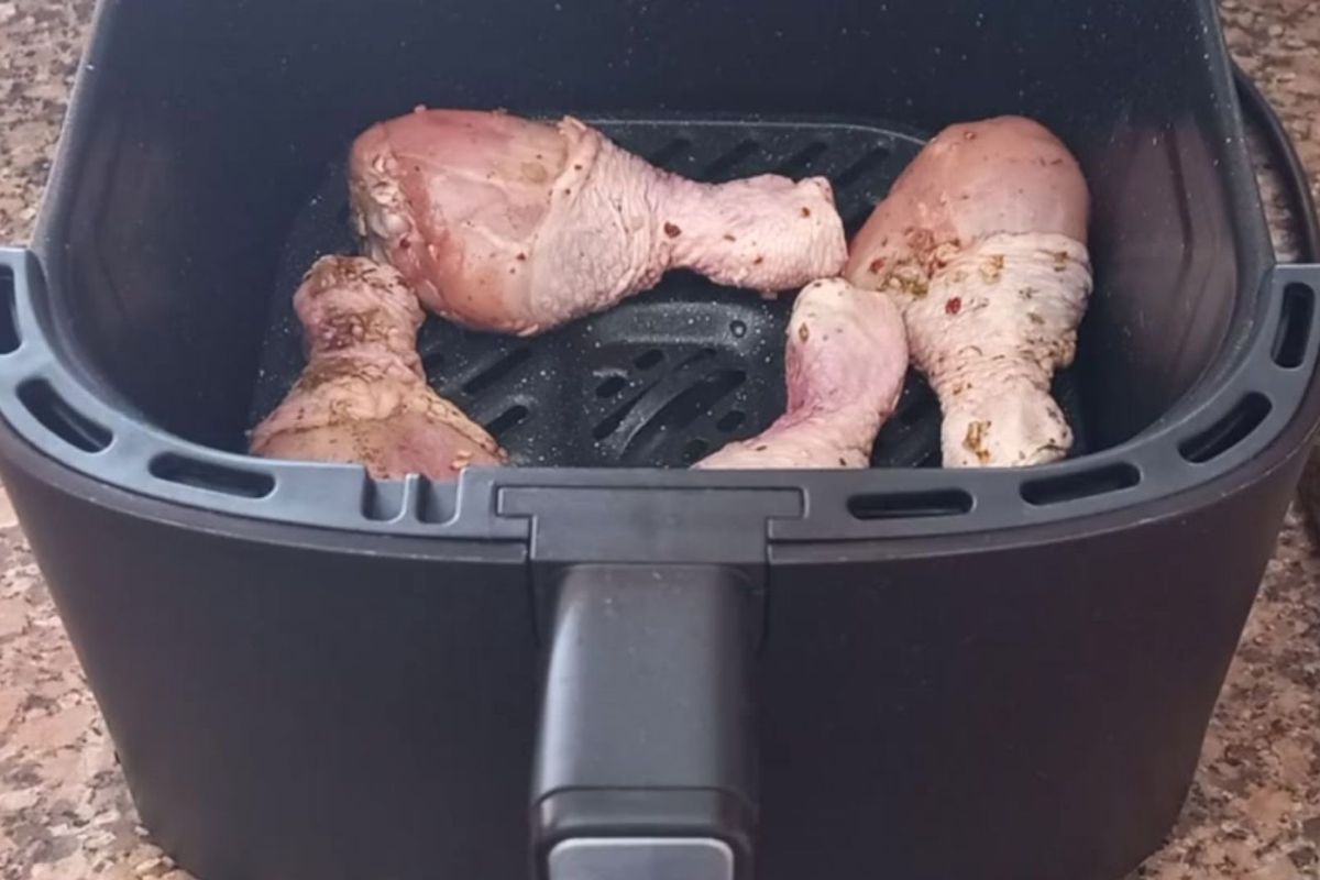 Quatro coxas de frango cruas e temperadas organizadas dentro do cesto de uma airfryer preta antes do cozimento